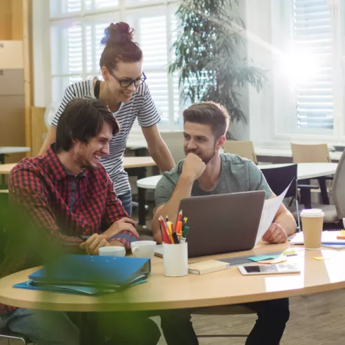 Three individuals working collaboratively around a single computer on a coffee table with colorful pencils, fostering a creative and relaxed atmosphere in an office setting